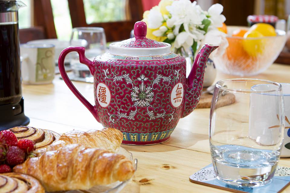 A maroon japanese style teapot on a wooden kitchen table with a croissant a a glass of water