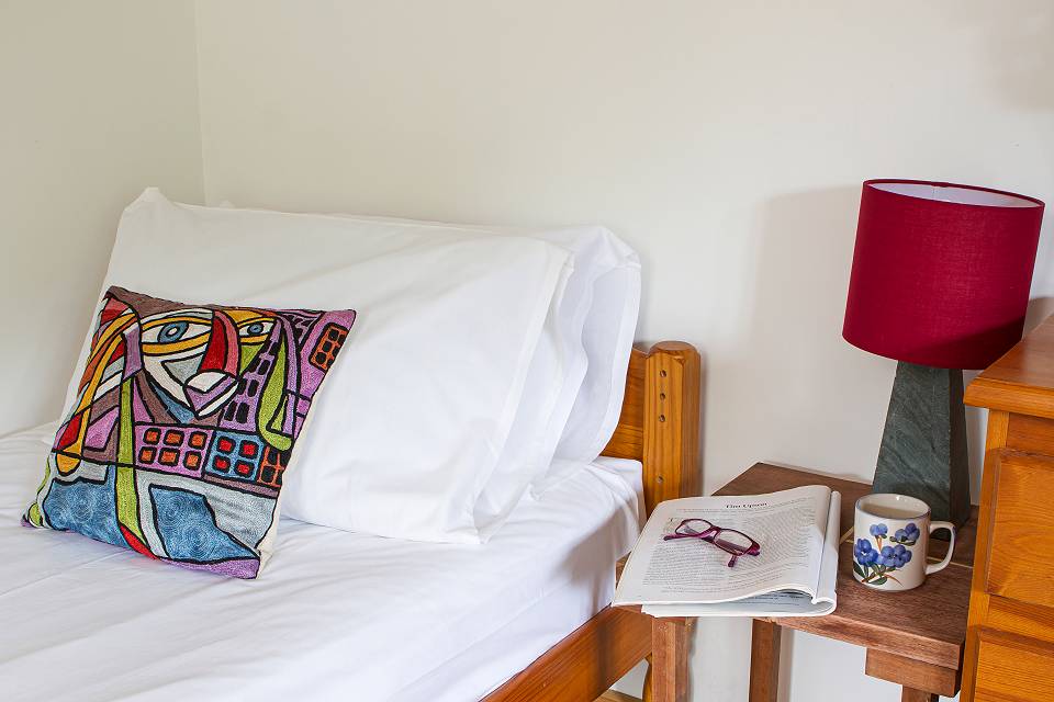 Close up of a bed with a colourful cushion. A cup of tea and a magazine and reading glasses on the bedside table